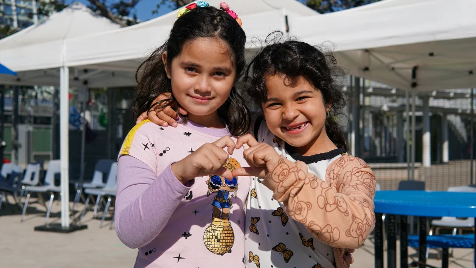 Two young girls making a heart sign with their fingers in front of a tent.