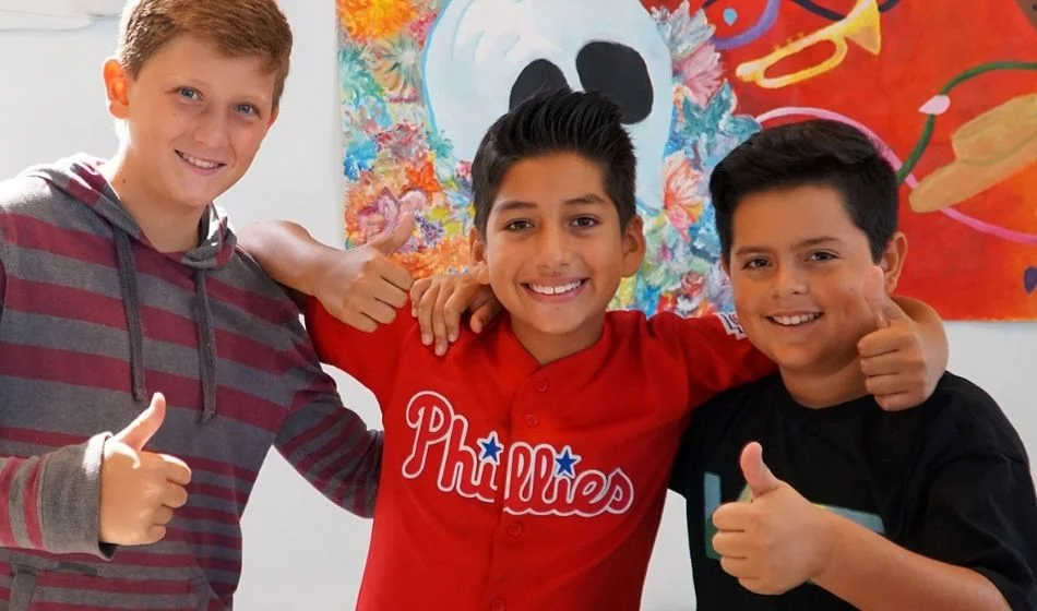 Three boys giving thumbs up in front of a painting.