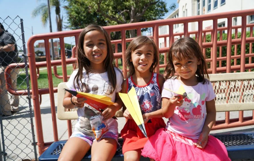 Three young girls hold paper airplanes while sitting on a bench