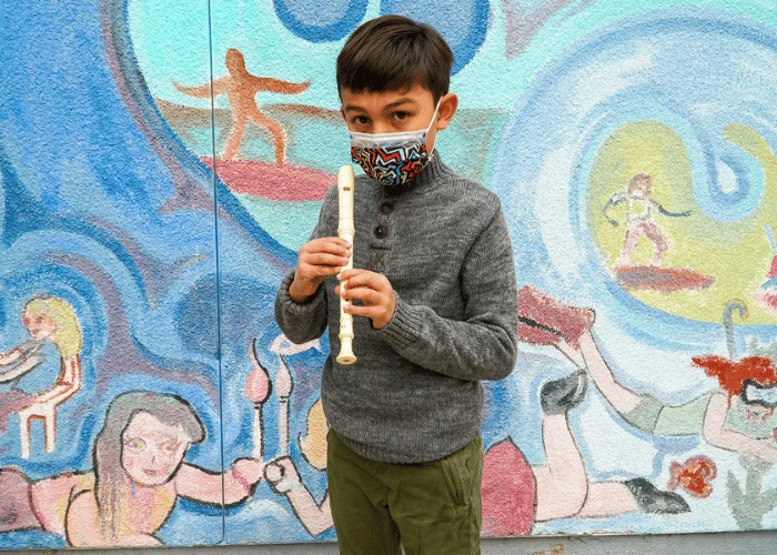 A boy wearing a mask and playing a flute in front of a mural.