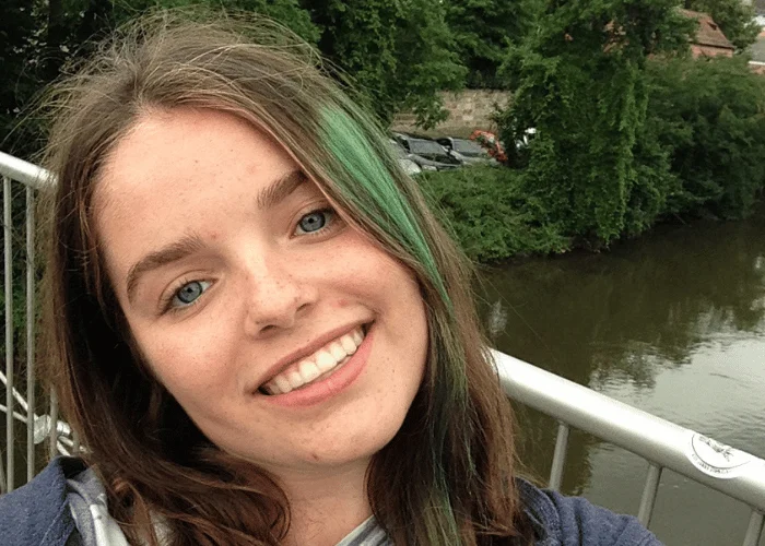 A woman with green in her hair standing on a bridge near a river.